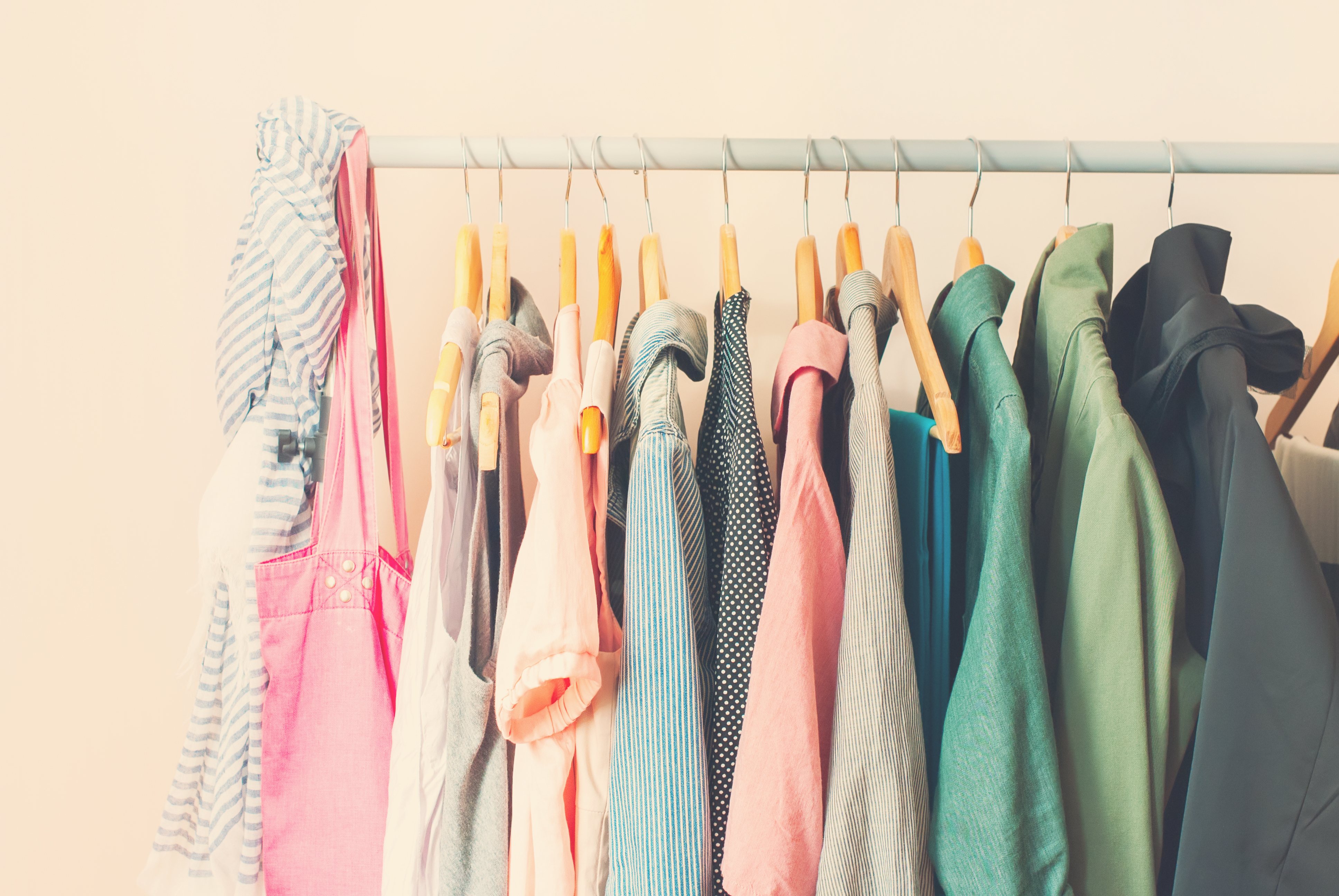 a rack of clothes with brightly colored shirts and a pink purse hanging off of it, with a pale tan background behind it