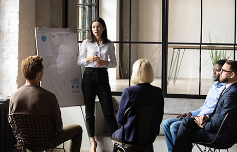 a group of professionals dressed in suits sit and listen to a facilitator who describes items on her whiteboard